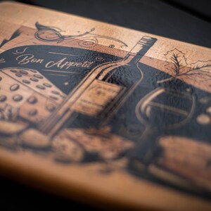 May include: A wooden cutting board with a black and white design featuring a bottle of wine, a glass of wine, a piece of cheese, and grapes. The text "Bon Appetit" is written on the board.