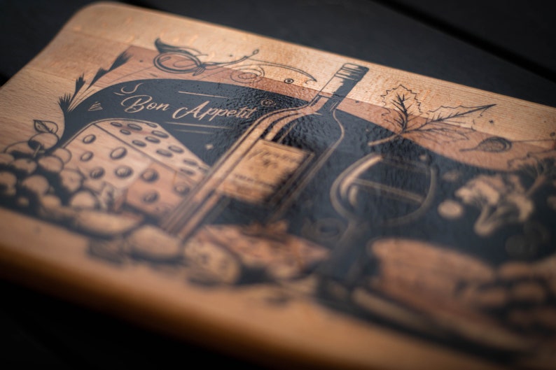 May include: A wooden cutting board with a black and white design featuring a bottle of wine, a glass of wine, a piece of cheese, and grapes. The text "Bon Appetit" is written on the board.