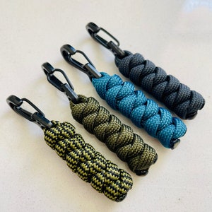 Könnte beinhalten: Vier Schlüsselanhänger mit schwarzen Metallclips und geflochtenem Paracord in Schwarz, Türkis und Olivgrün.