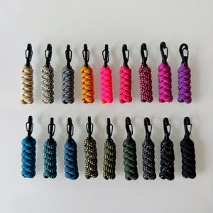 Könnte beinhalten: Achtzehn Paracord-Reißverschlussanhänger in zwei Reihen, jeweils mit einer geflochtenen Kordel in verschiedenen Farben und Mustern an einem schwarzen Kunststoff-Karabinerhaken. Die Farben umfassen Beige, Grau, Schwarz, Orange, Pink, Lila, Blau und Grün.