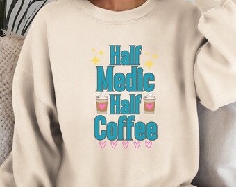 Op de afbeelding: Een beige sweatshirt met de tekst "Half Medic Half Coffee" in blauw met twee koffiebekers en roze hartjes.