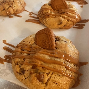 May include: Close-up of a large, golden brown cookie topped with caramel sauce and a single, square-shaped cookie. The cookie is on a white parchment-lined baking sheet.