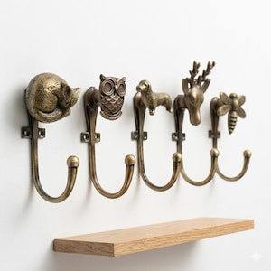 May include: Five antique brass wall hooks, each featuring a different animal: a cat, an owl, a dog, a deer, and a bee. They are mounted on a white wall above a wooden shelf, creating a decorative storage solution.