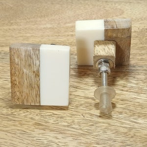 May include: Two square wooden cabinet knobs with a white resin top half. The knobs have a silver screw and a clear plastic base.