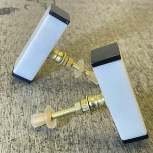 Puede incluir: Dos pomos de armario de cerámica blanca con bases cuadradas negras y herrajes metálicos dorados.