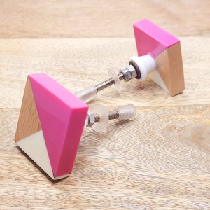 May include: Two pink and white geometric cabinet knobs with a wood accent. The knobs have a white center and a pink outer edge. The knobs are attached to a silver metal screw.
