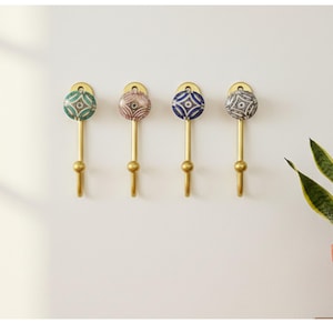 May include: Four decorative wall hooks with brass stems and ceramic tops. The ceramic tops display geometric patterns in teal, pink, blue, and grey. The hooks are mounted on a white wall, providing a stylish storage solution for any room.