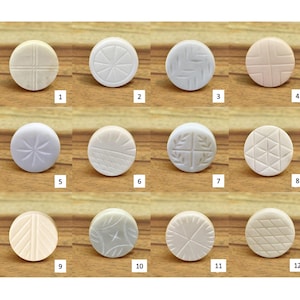 May include: Twelve round, off-white decorative buttons with carved designs. Patterns include crosshatch, starburst, and geometric shapes. The buttons are displayed on a wooden surface. Each button has a diameter of approximately 2.5 cm.