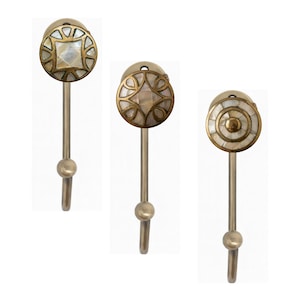 May include: Three decorative brass hooks with intricate designs. Each hook features a round, decorative head with a mother-of-pearl inlay and a brass stem. The designs include a square, a diamond, and a circular pattern. The hooks are ideal for hanging items.