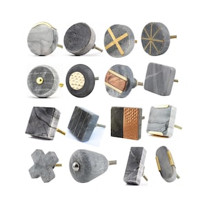 Könnte beinhalten: Eine Sammlung dekorativer Schrankknöpfe in verschiedenen Formen und Designs. Die Knöpfe sind aus grauem Stein gefertigt und weisen goldene und kupferfarbene Akzente auf. Zu den Stilen gehören runde, quadratische und kreuzförmige Formen mit unterschiedlichen Oberflächenstrukturen.