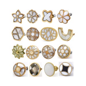 May include: A collection of decorative cabinet knobs in various shapes, including stars, flowers, and geometric designs. Each knob features a gold-colored metal base and is inlaid with white mother-of-pearl. The knobs are designed for furniture and home decor.