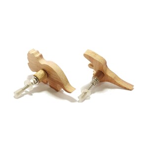 May include: Two wooden dinosaur-shaped cabinet knobs. The knobs are light brown with a natural wood grain finish. Each knob has a metal screw and a clear plastic washer for installation. The knobs are designed for furniture.