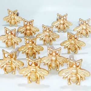 Może przedstawiać: Zestaw 12 złotych gałek do szuflad w kształcie pszczół. Gałki wykonane są z metalu i mają błyszczące wykończenie. Każda gałka ma małą pętlę z tyłu do mocowania do szuflady.