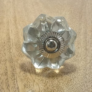 Könnte beinhalten: Ein klarer Glas-Schrankknopf mit einem silbernen Metallzentrum. Der Knopf hat eine blumenartige Form mit einem facettierten Design.