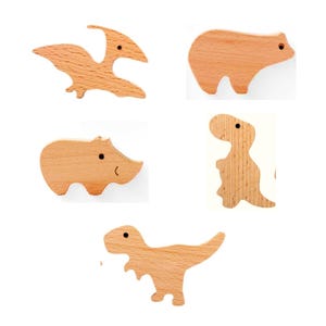 May include: Five wooden animal cutouts: a pterodactyl, bear, hippo, dinosaur, and T-Rex. Each cutout is made of light-colored wood with a smooth finish. The bear and hippo have simple, rounded shapes, while the dinosaurs have more detailed silhouettes.