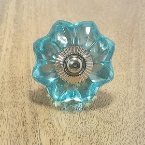 May include: A clear, turquoise glass cabinet knob with a flower-like design. The knob has a silver-colored metal base and a rounded center. The glass is faceted, creating a textured appearance. This decorative hardware is suitable for furniture.