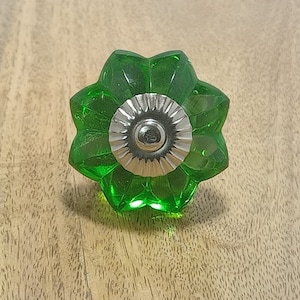 May include: A bright green, flower-shaped cabinet knob with a silver-colored metal base. The knob is made of transparent glass and sits on a light brown wooden surface. The knob's design features eight petals and a central screw fitting.