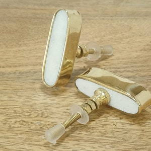 Könnte beinhalten: Zwei ovale Schrankknöpfe mit einem weißen Keramikkern und goldfarbener Metallverkleidung. Die Knöpfe haben ein Schraubdesign.