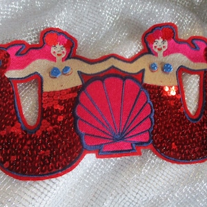 May include: Two red sequin mermaid figures with pink hair and blue seashells on their chests, flanking a pink seashell with a blue outline.
