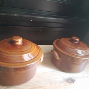 May include: Two brown ceramic casserole dishes with lids. The dishes are on a wooden surface.