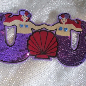 May include: A whimsical patch featuring two mermaids with red hair, holding hands above a red seashell. The mermaids have purple sequined tails and blue sequin accents. The patch has a purple border.