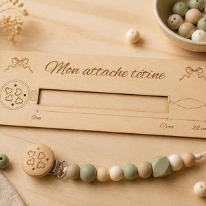 Peut inclure: Une planche de présentation en bois clair avec le texte Mon attache tétine, une règle graduée de 0cm à 22cm, et une attache-tétine composée de perles en silicone vert sauge, beige et blanc, un clip en bois gravé de cœurs et une boucle.