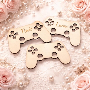 Puede incluir: Tres recortes de madera con forma de mando de videojuegos, con los nombres "Timéo" y "Lucas" grabados. Los mandos son de color marrón claro y tienen detalles de botones y joystick. El fondo incluye perlas y rosas rosas.
