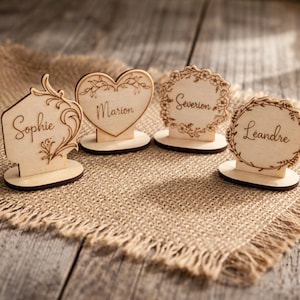 May include: Four wooden place card holders with names engraved on them. The holders are in various shapes: a house, a heart, and two circles. The names are "Sophie", "Marion", "Severion", and "Leandre". The holders are on a burlap surface.