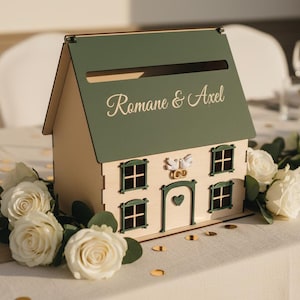 Könnte beinhalten: Eine holzfarbene, hausförmige Kartenbox mit grünem Dach und den Namen "Romane & Axel" in Schreibschrift. Die Box hat Fenster und eine herzförmige Tür. Weiße Rosen und Grünzeug schmücken den Tisch.
