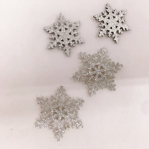 May include: Four silver glitter snowflake decorations. Each snowflake has six points and a detailed design. The decorations are scattered on a white surface, creating a festive arrangement. Suitable for holiday crafts and winter decor.