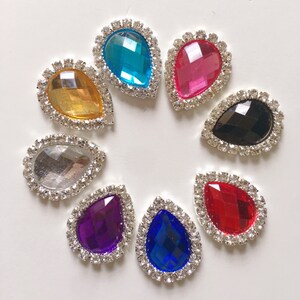 Puede incluir: Una colección de ocho adornos de pedrería en forma de lágrima en varios colores, incluyendo amarillo, turquesa, rosa, negro, plateado, morado, azul y rojo. Cada gema está rodeada por un borde plateado con incrustaciones de pedrería.