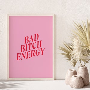 Puede incluir: Un p&oacute;ster rosa enmarcado con el texto "BAD BITCH ENERGY" en letras rojas. El p&oacute;ster est&aacute; colgado en una pared blanca con una hoja de palma seca y hierba de la pampa en un jarr&oacute;n en primer plano.