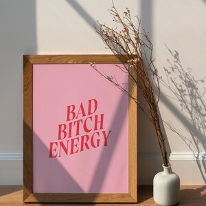 Puede incluir: Un p&oacute;ster rosa enmarcado en madera con el texto "BAD BITCH ENERGY" en letras rojas. El p&oacute;ster est&aacute; apoyado en una pared blanca con un arreglo de flores secas en un jarr&oacute;n blanco.