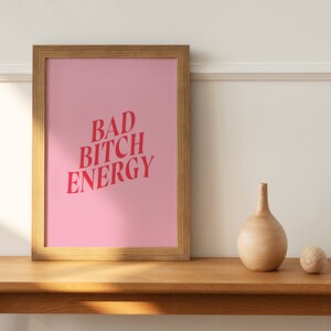 Puede incluir: Una impresi&oacute;n enmarcada con un fondo rosa y el texto "BAD BITCH ENERGY" en letras rojas. La impresi&oacute;n est&aacute; sobre una mesa de madera con un jarr&oacute;n de madera y una bola de madera.