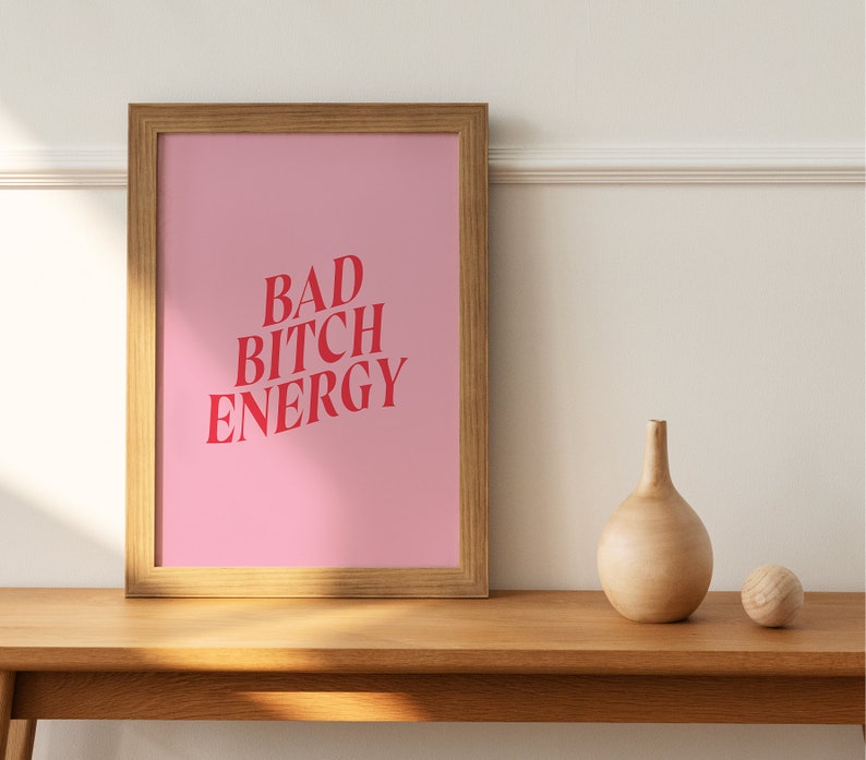 Puede incluir: Una impresi&oacute;n enmarcada con un fondo rosa y el texto "BAD BITCH ENERGY" en letras rojas. La impresi&oacute;n est&aacute; sobre una mesa de madera con un jarr&oacute;n de madera y una bola de madera.