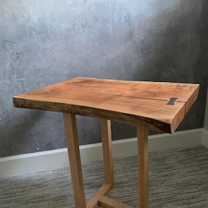 May include: A handcrafted wooden side table with a natural edge. The tabletop is a warm brown color, supported by light-colored wooden legs. The table is approximately 30 inches tall.