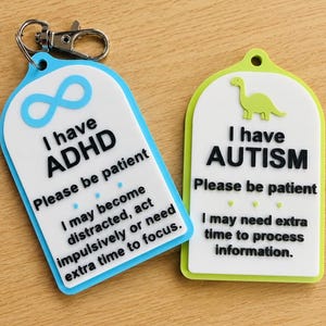 May include: Two rectangular silicone tags with rounded tops. The blue tag features a light blue infinity symbol, the text I have ADHD, Please be patient, and I may become distracted, act impulsively or need extra time to focus. The lime green tag features a green dinosaur, the text I have AUTISM, Please be patient, and I may need extra time to process information. The blue tag includes a metal lobster clasp.