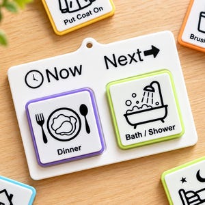 May include: A white plastic visual schedule board with a Now and Next section. The Now section features a purple-bordered tile with a dinner icon, while the Next section has a green-bordered tile with a bath and shower icon.