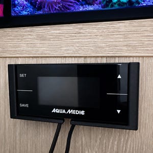May include: A black digital controller with the text "AQUA MEDIC" and control buttons. The controller is mounted on a light wood-grain surface. The display shows a blank screen. The words "SET" and "SAVE" are visible.