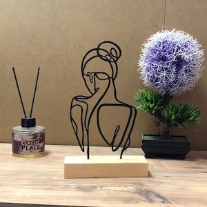 May include: A black metal sculpture of a woman's silhouette, mounted on a light wood base. The artwork is minimalist and modern, with a focus on the lines of the figure. The sculpture is displayed on a wooden surface, alongside a decorative plant and a fragrance diffuser.