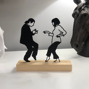 May include: Black metal silhouette of a dancing couple on a wooden base. The figures are stylized, with the man in a suit and the woman in a blouse and trousers. The sculpture is a decorative item.
