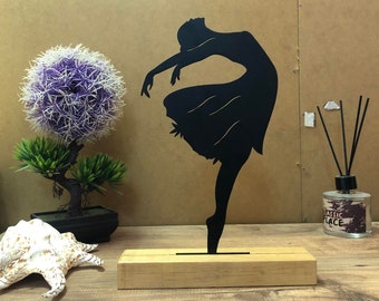 May include: Black metal silhouette of a dancer in a flowing skirt, standing on a wooden base. The sculpture is on a wooden table with a potted plant and a decorative seashell.