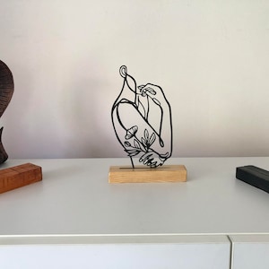 May include: Black metal sculpture of a person with floral details, standing on a wooden base. The sculpture is on a white surface.