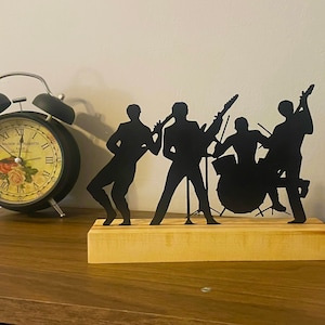 Könnte beinhalten: Schwarze Metallsilhouette einer Band, die Musik spielt. Die Band besteht aus einem Schlagzeuger, einem Gitarristen, einem Bassisten und einem Saxophonisten. Die Silhouette ist auf einem Holzsockel montiert.