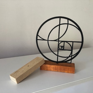 May include: A black metal sculpture of a geometric design, resembling a spiral, on a wooden base. The sculpture is on a white surface.