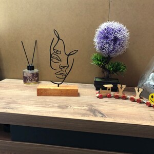 May include: Black metal wire sculpture of two faces on a wooden base. The sculpture is on a wooden table with a small potted plant and a toy train in the background.