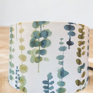May include: A white lampshade featuring a watercolour pattern of green and blue leaves. The lampshade is made from fabric and has a cylindrical shape.