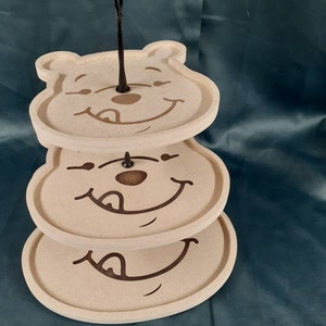 May include: A three-tiered serving tray featuring Winnie the Pooh's face on each tier. The tray is made of a light beige material and has a black metal handle.