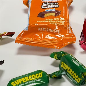 May include: An orange Choco Cake Mini candy wrapper with text and a picture of the cake. The wrapper states "Golden cake with chocolate flavor coating". Also visible are green Supercoco candy wrappers.