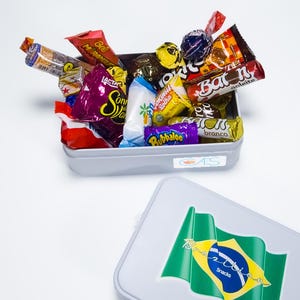 Pode incluir: Uma lata retangular cinza cheia de vários doces embalados. A lata está transbordando de guloseimas coloridas, incluindo barras de chocolate e pirulitos. Uma segunda lata com a bandeira brasileira e as palavras "Brazilian Snacks" está em primeiro plano.